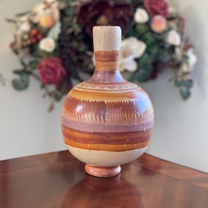 May include: A handcrafted, round ceramic vase with a wide base and a narrow neck. The vase features horizontal bands of color in shades of tan, orange, and purple. The vase is displayed on a wooden surface.