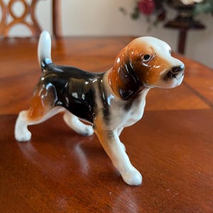 May include: A ceramic beagle dog figurine with a glossy finish. The dog has a white, brown, and black coat, with a white tail and paws. The figurine is posed in a standing position, with its head turned slightly to the right.