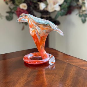 May include: A vibrant, hand-blown glass sculpture in the shape of a cobra. The art glass piece features swirling patterns of orange, red, and blue. The base is a coiled ring, and the head flares out. The sculpture is displayed on a wooden surface.