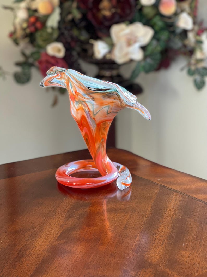 May include: A vibrant, hand-blown glass sculpture in the shape of a cobra. The art glass piece features swirling patterns of orange, red, and blue. The base is a coiled ring, and the head flares out. The sculpture is displayed on a wooden surface.