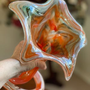 May include: A hand-blown glass art piece with a unique, organic shape. The glass features swirls of orange, white, and blue, creating a vibrant and dynamic visual effect. The piece has a flared, open top and a curved, stem-like base.