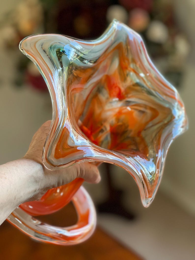 May include: A hand-blown glass art piece with a unique, organic shape. The glass features swirls of orange, white, and blue, creating a vibrant and dynamic visual effect. The piece has a flared, open top and a curved, stem-like base.