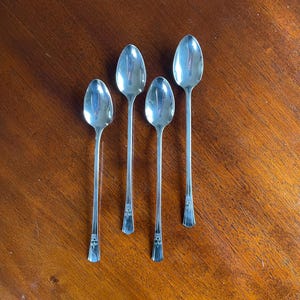 May include: Four silver spoons arranged on a dark wooden surface. The spoons have a classic design with elongated handles and decorative detailing at the base. The spoons are reflective, showing the surrounding environment.