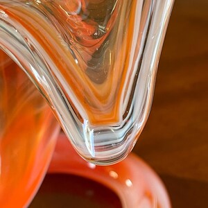 May include: Close-up of a blown glass sculpture with a swirling pattern of orange, white, and clear glass. The glass has a glossy finish and a unique, organic shape. The sculpture's design features a blend of colors and textures.