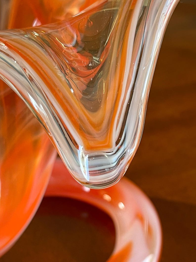 May include: Close-up of a blown glass sculpture with a swirling pattern of orange, white, and clear glass. The glass has a glossy finish and a unique, organic shape. The sculpture's design features a blend of colors and textures.