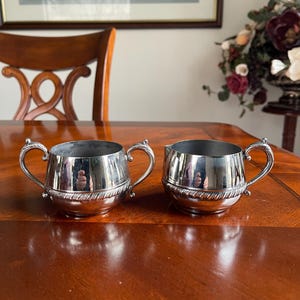 May include: Two silver-plated serving bowls with handles. The bowls have a rounded shape with a decorative rope-like design around the middle. They are sitting on a polished wooden surface. The background includes a wooden chair, a framed picture, and a floral arrangement.
