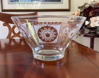 May include: Clear glass bowl with a flared design. The bowl features a brown and white decorative pattern with a crown motif. The bowl is sitting on a wooden table. The bowl is likely vintage.