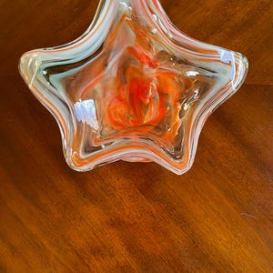 May include: A star-shaped glass bowl with a vibrant orange, white, and light blue swirl design. The bowl is transparent, allowing the colors to blend and create a unique pattern. The bowl is a decorative piece.