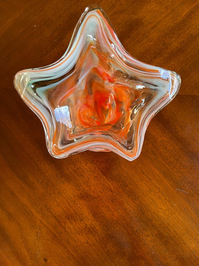 May include: A star-shaped glass bowl with a vibrant orange, white, and light blue swirl design. The bowl is transparent, allowing the colors to blend and create a unique pattern. The bowl is a decorative piece.