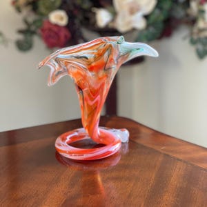 May include: A vibrant, hand-blown glass vase with a unique, swirling design. The vase features a bright orange, white, and black color scheme, with a flared top and a coiled base. The vase is a decorative art piece.