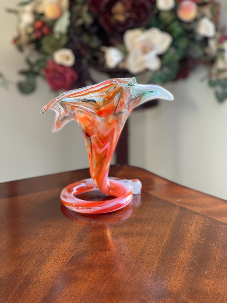 May include: A vibrant, hand-blown glass vase with a unique, swirling design. The vase features a bright orange, white, and black color scheme, with a flared top and a coiled base. The vase is a decorative art piece.