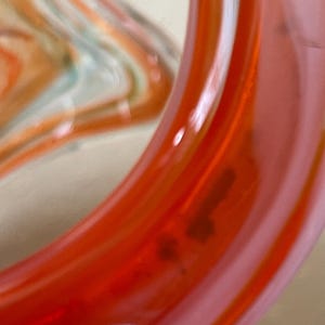 May include: Close-up of a section of a glass object with a vibrant orange and pink color scheme. The glass has a smooth, glossy finish with a swirl design. The object appears to be a decorative piece or part of a larger art installation.