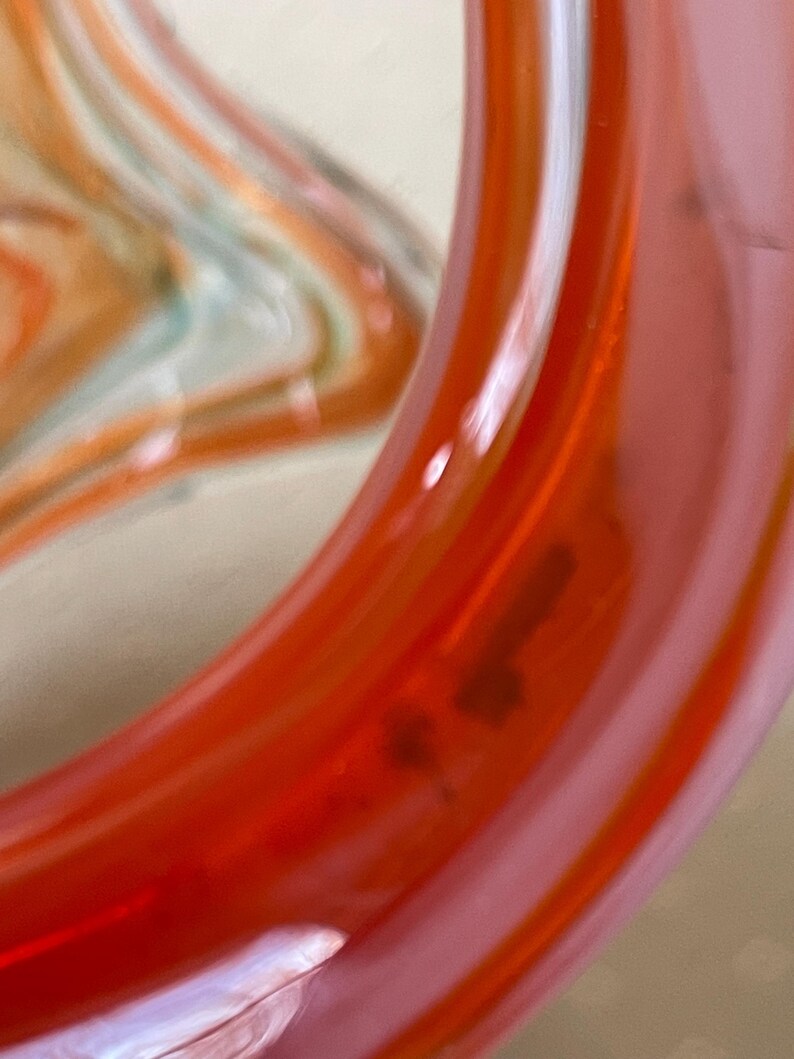 May include: Close-up of a section of a glass object with a vibrant orange and pink color scheme. The glass has a smooth, glossy finish with a swirl design. The object appears to be a decorative piece or part of a larger art installation.