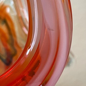 May include: Close-up of a section of a glass object with a rounded shape. The glass is a gradient of red and pink, with a crack visible on the surface. The interior of the glass shows swirls of orange and white.