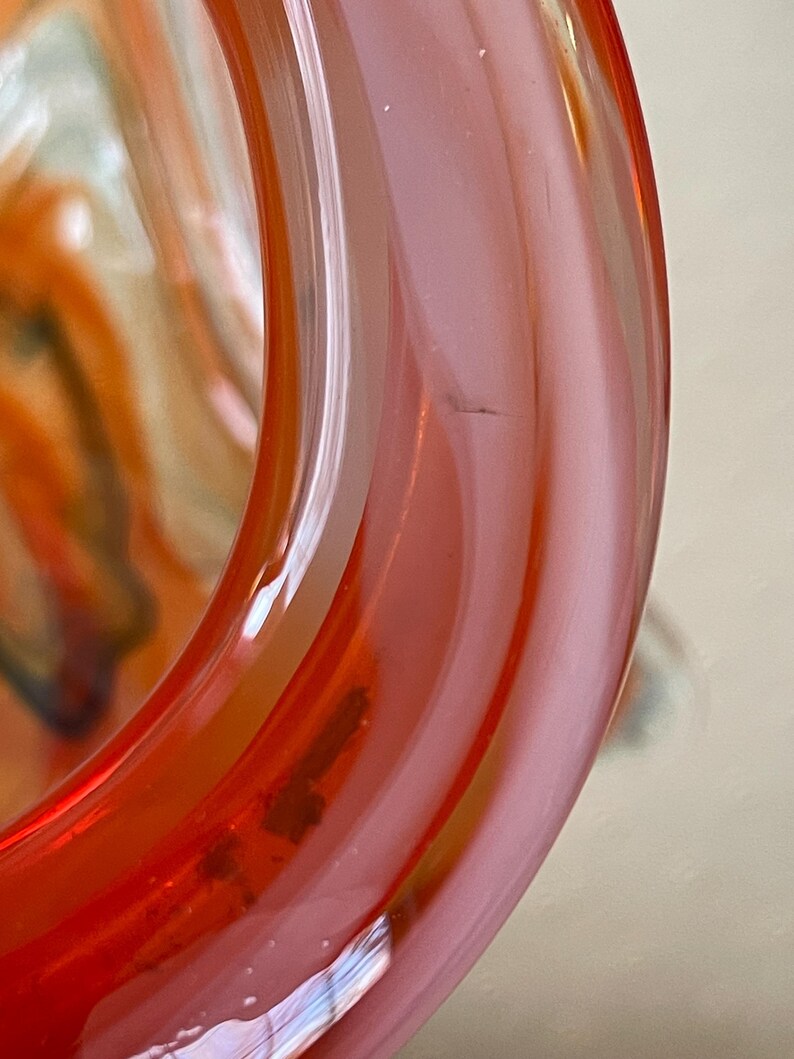 May include: Close-up of a section of a glass object with a rounded shape. The glass is a gradient of red and pink, with a crack visible on the surface. The interior of the glass shows swirls of orange and white.