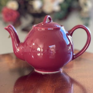 May include: A claret-coloured ceramic teapot with a rounded body, a curved spout, and a loop handle. The lid has a small handle. The teapot is on a wooden surface, with a blurred floral background.