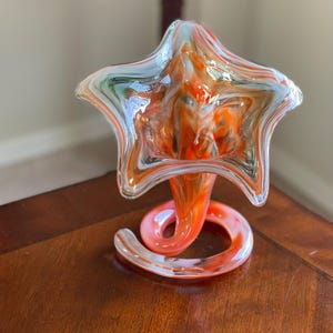 May include: A decorative glass vase with a star-shaped top and a swirling, spiral base. The vase features a blend of orange, white, and light blue colors. The vase is sitting on a wooden surface.