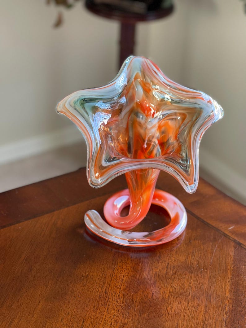 May include: A decorative glass vase with a star-shaped top and a swirling, spiral base. The vase features a blend of orange, white, and light blue colors. The vase is sitting on a wooden surface.