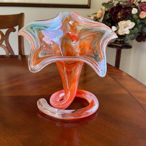 May include: A vibrant, hand-blown glass vase with a unique design. The vase features a flared, star-shaped top in shades of orange, white, and blue. The stem is a spiraling shape, adding a touch of elegance. A decorative piece for any home.