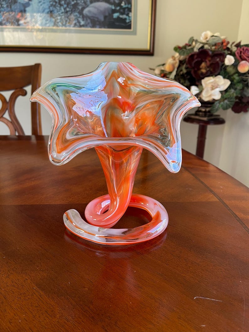 May include: A vibrant, hand-blown glass vase with a unique design. The vase features a flared, star-shaped top in shades of orange, white, and blue. The stem is a spiraling shape, adding a touch of elegance. A decorative piece for any home.