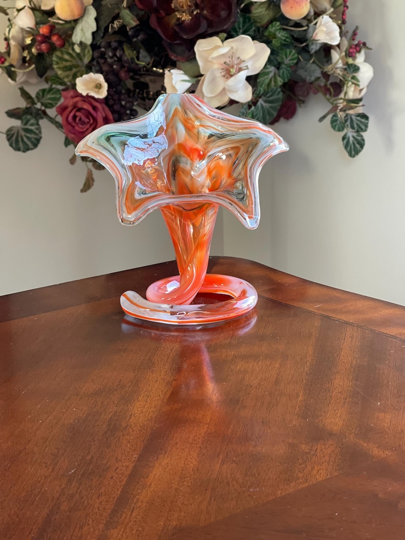 May include: A vibrant, hand-blown glass vase with a star-shaped top and a flared base. The vase features a swirl of orange, white, and blue colors. The base has a decorative, curled design. The vase is approximately 10 inches tall.