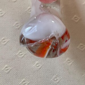 May include: A clear glass marble with a white and orange swirl design. The marble has a rounded shape and is partially transparent, revealing the colorful interior. The top portion is white, while the bottom features orange and white stripes.