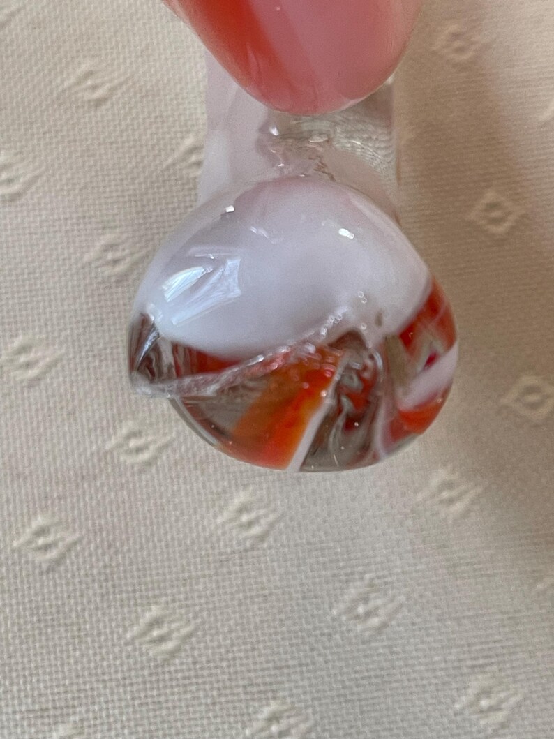 May include: A clear glass marble with a white and orange swirl design. The marble has a rounded shape and is partially transparent, revealing the colorful interior. The top portion is white, while the bottom features orange and white stripes.