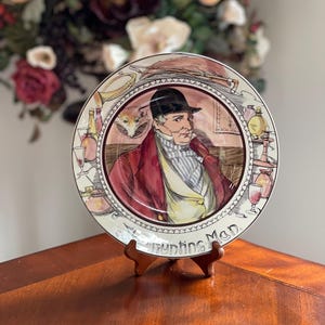 May include: Decorative plate featuring a man in a red coat and black hat, with the text "Hunting Man." The plate has a detailed illustration with a fox and bottles. It is displayed on a wooden stand.