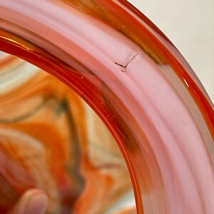 May include: Close-up of a glass bowl with a vibrant swirl design. The bowl features shades of orange, pink, and a hint of green. The rim is a solid, translucent orange. A small crack is visible on the rim.
