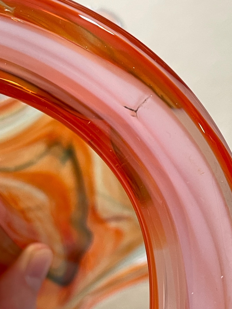 May include: Close-up of a glass bowl with a vibrant swirl design. The bowl features shades of orange, pink, and a hint of green. The rim is a solid, translucent orange. A small crack is visible on the rim.