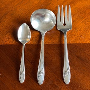 May include: A silver spoon, ladle, and fork set on a wooden surface. The utensils have a floral design on the handles. The spoon is small, the ladle is round, and the fork has three tines.