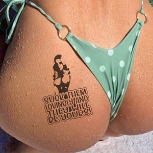 May include: A close-up of a person's skin with a temporary tattoo and a green polka dot bikini bottom. The tattoo features a silhouette of a person and the text "SUCK THEM LOVINGLY AND THEY WILL BE YOURS!"