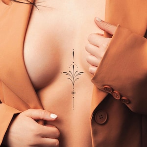 May include: A black, symmetrical tattoo design on the chest featuring a central vertical line with floral and dot details. The design is centered and framed by a tan blazer.