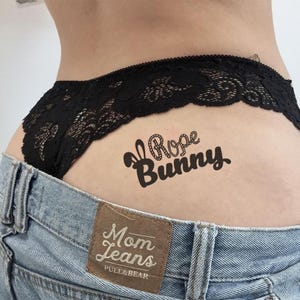 Może przedstawiać: Czarne koronkowe majtki z ozdobnym brzegiem, połączone z jasnymi dżinsami. Tymczasowy tatuaż z napisem "Rose Bunny" jest widoczny na skórze. Dżinsy mają brązową skórzaną naszywkę z napisem "Mom Jeans" i marką "PULL&BEAR".