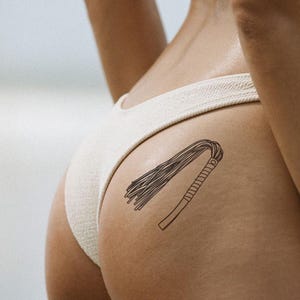 Puede incluir: Un tatuaje temporal de un diseño de látigo negro. El tatuaje muestra un mango con un agarre texturizado y un conjunto de hebras finas. El diseño está ubicado en la parte baja de la espalda.