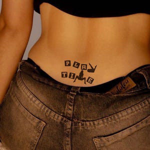 May include: Black tattoo on the lower back that reads "PLAY TIME" with a silhouette of a woman. The tattoo is above the waistband of a pair of faded denim jeans.