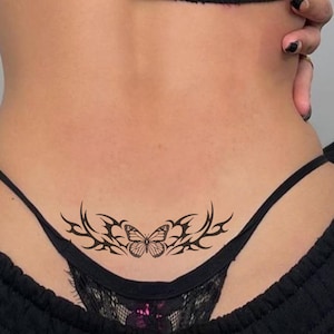 Puede incluir: Diseño de tatuaje de mariposa negra en la parte baja de la espalda. El tatuaje presenta una mariposa detallada en el centro, flanqueada por patrones de estilo tribal. El diseño está sobre la piel, con un tanga negro visible.