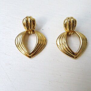 May include: A pair of gold-tone dangle earrings with a heart-shaped design. The earrings have a textured, ribbed surface and a small loop at the top.