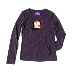 May include: A long-sleeved, dark purple shirt with a crew neck. The shirt features a square patch on the chest with floral embroidery and a small rectangular patch near the neckline. The fabric has a subtle heathered texture.