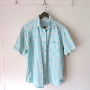 May include: A short-sleeved, button-down shirt in a light blue and cream plaid pattern. The shirt has a single chest pocket and is hanging on a wooden hanger against a white wall. The shirt is made of a lightweight fabric.