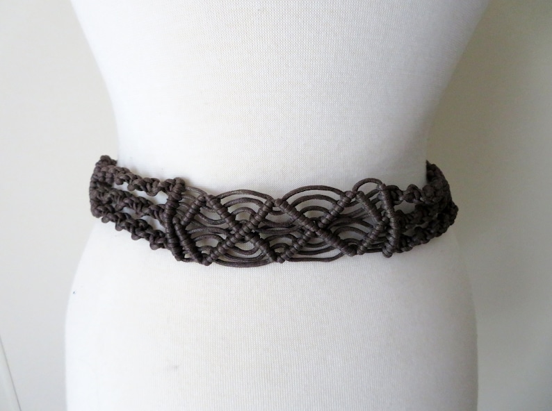 Puede incluir: Un cintur&oacute;n de macram&eacute; marr&oacute;n con un dise&ntilde;o tejido. El cintur&oacute;n se muestra en un maniqu&iacute; blanco, mostrando sus intrincados nudos y estilo general. El cintur&oacute;n tiene aproximadamente 5 cm de ancho.