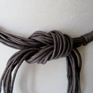 Puede incluir: Primer plano de un collar marr&oacute;n con m&uacute;ltiples hebras anudadas. El collar parece estar hecho de un material tejido, y el nudo est&aacute; centrado en el marco. El fondo es una tela lisa de color blanquecino.