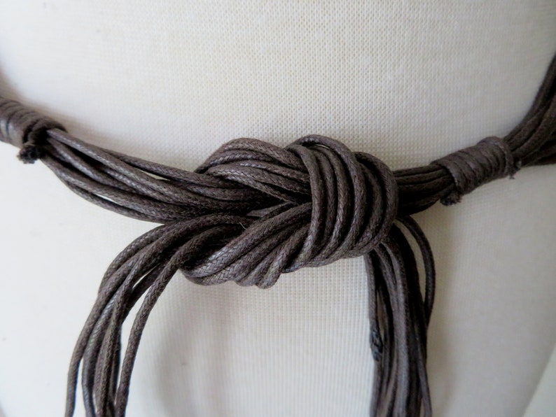 Puede incluir: Primer plano de un collar marr&oacute;n con m&uacute;ltiples hebras anudadas. El collar parece estar hecho de un material tejido, y el nudo est&aacute; centrado en el marco. El fondo es una tela lisa de color blanquecino.