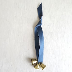 May include: A decorative hanging ornament featuring three small, gold-colored bells suspended from a knotted, blue ribbon. The ribbon is a solid, muted blue color. The bells are clustered together at the bottom of the ribbon.