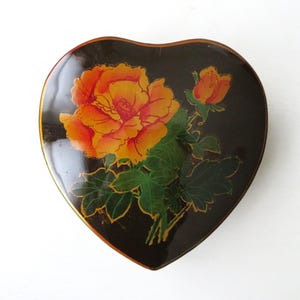 Puede incluir: Caja de baratijas en forma de corazón con un acabado negro brillante. La tapa presenta un diseño floral vibrante con rosas naranjas y amarillas, hojas verdes y detalles dorados. Un artículo de almacenamiento decorativo.