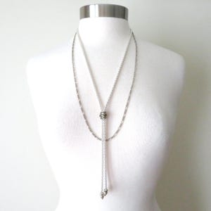 May include: A silver-toned lariat necklace featuring a long, delicate chain that loops through a central metallic ring. The chain ends in two small, dangling silver-toned charms, creating a layered effect against the white display form.