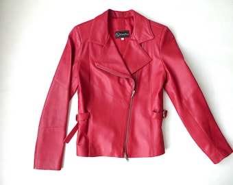 May include: A vibrant red leather jacket featuring an asymmetrical front zipper closure, a wide notched collar, and long sleeves. The jacket includes adjustable side buckle straps at the waist and a black interior brand label near the neckline.