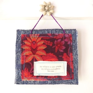 May include: A square fabric wall hanging featuring a dark blue denim-like border and a central panel with a vibrant red and orange floral pattern. A white rectangular fabric patch is stitched on the front with the text: my religion is very simple my religion is kindness Dalai Lama. It hangs from a purple string attached to a white flower-shaped hook.