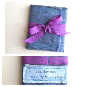 May include: A blue fabric cover with a subtle polka dot pattern, frayed edges, and a purple ribbon tied in a bow. The lower section features a rectangular fabric patch with the text "Don't bother me. I'm in my happy place." stitched onto the surface.