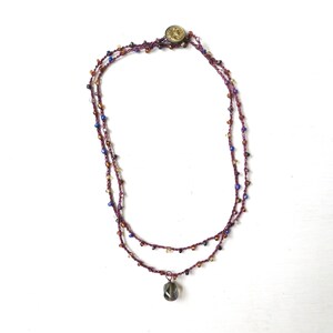 May include: A handcrafted necklace featuring two strands of dark purple thread adorned with small, colorful beads. A faceted, gray pendant hangs from the lower strand. The necklace is fastened with a gold-colored button.